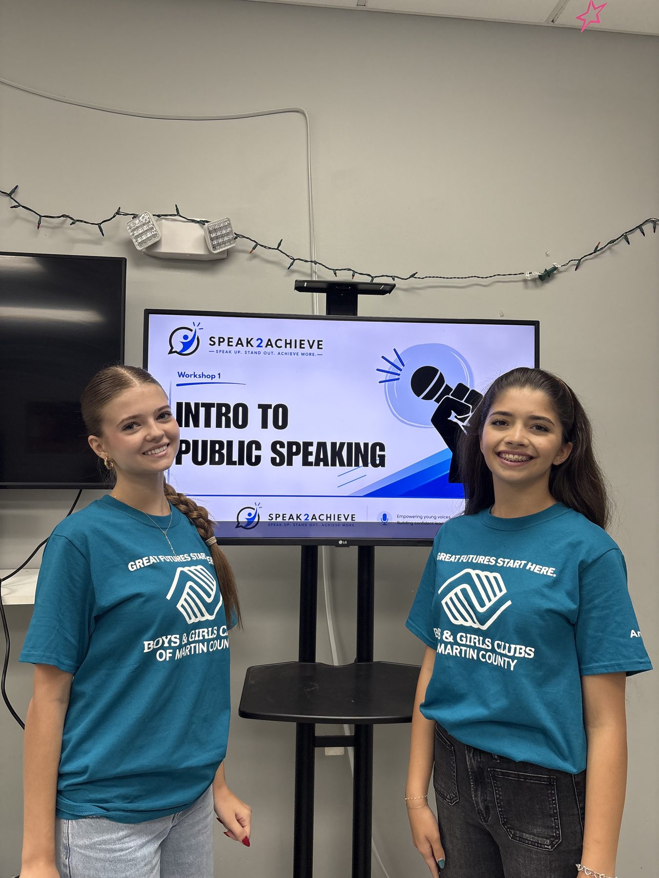 Two Speak2Achieve volunteers standing beside a screen showing the Intro to Public Speaking workshop slide
